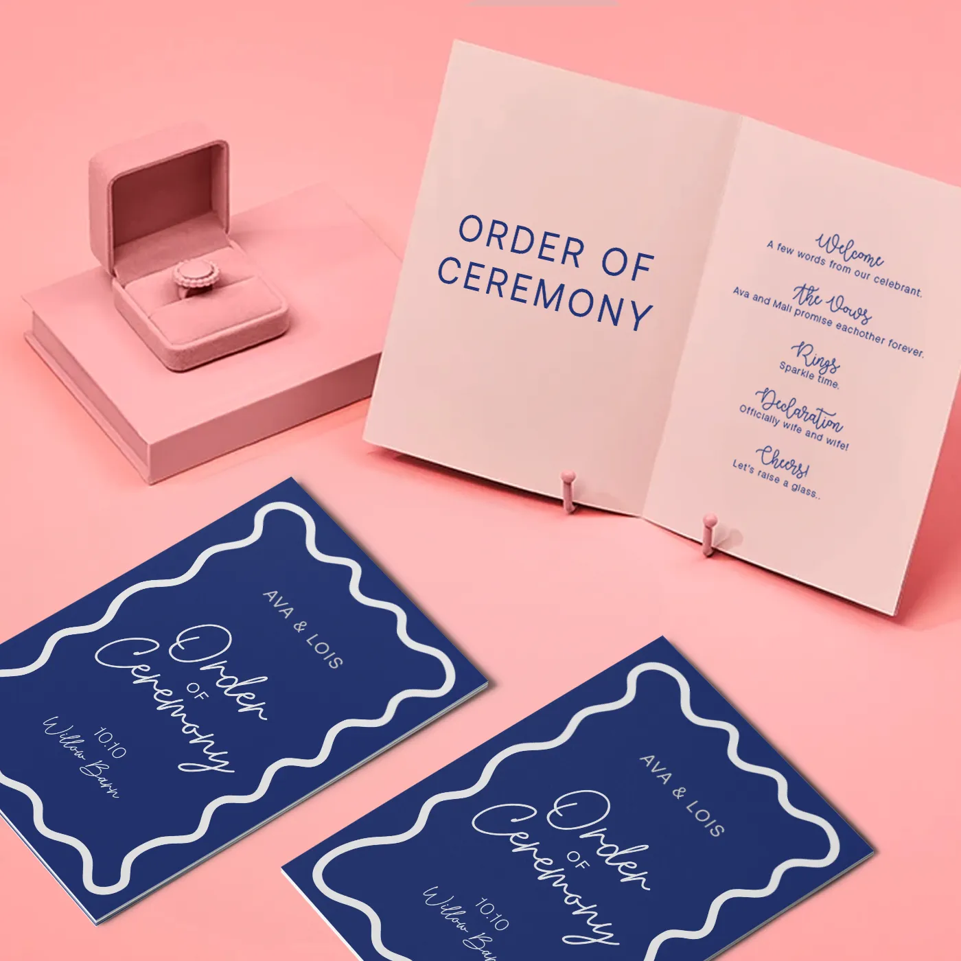Gemini said Two navy blue, flat-folded wedding ceremony programs for Ava and Lois featuring a white wavy border are shown on a light pink surface next to an open pink booklet that details the "Order of Ceremony," including "The Vows" and "Declaration." A pink velvet ring box sits nearby, holding a matching pink ring.