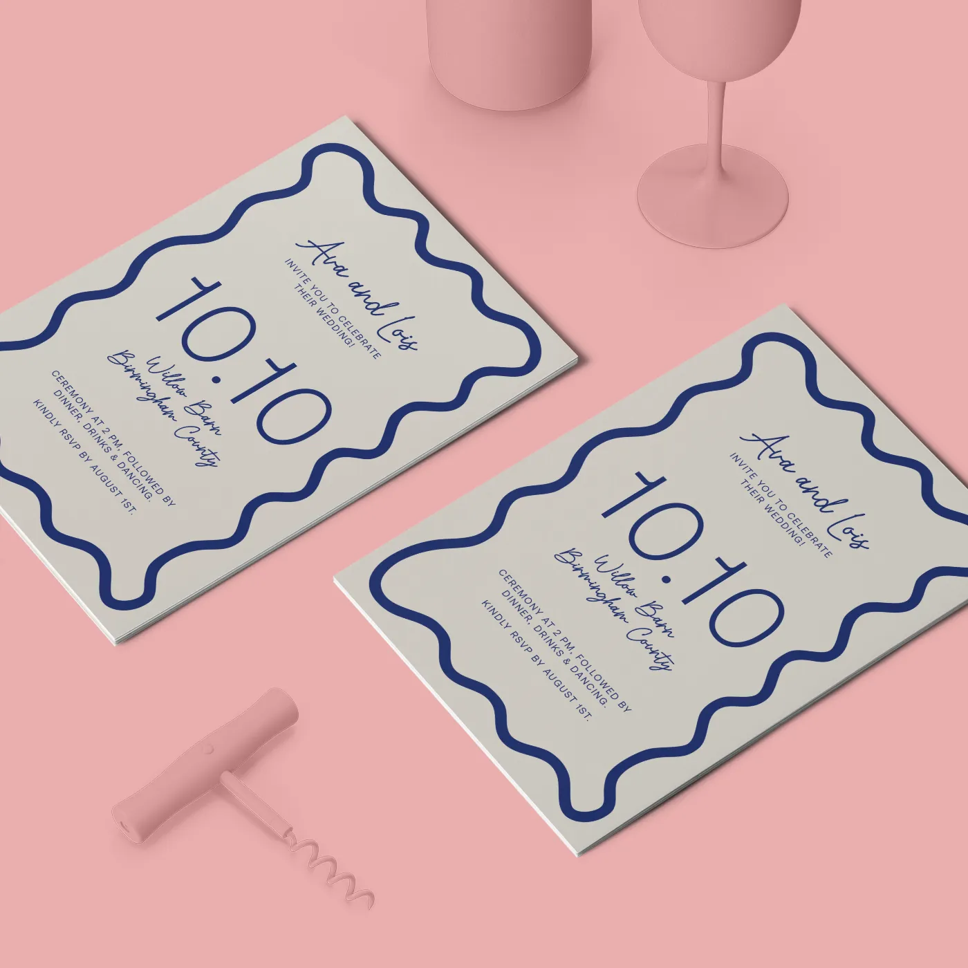 Two identical flat wedding invitations for "Ava and Lois" feature a navy blue wavy border and script font. They are displayed on a light pink surface next to pink-toned props, including two wine glasses and a wine bottle opener.