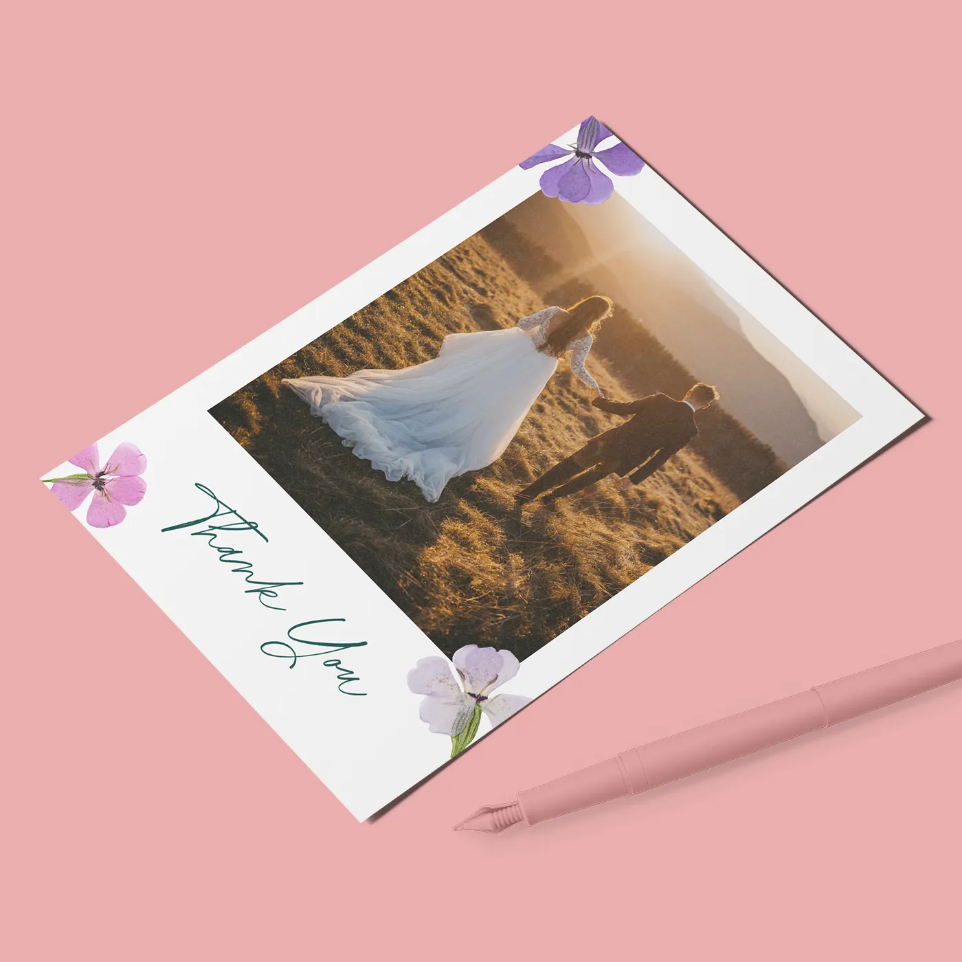 A white wedding thank-you card features a large central photo of a couple in a field at sunset, accented with four purple and pink pressed flowers in the corners and the words "Thank You" in green script. The card is shown on a light pink background next to a matching pink fountain pen.