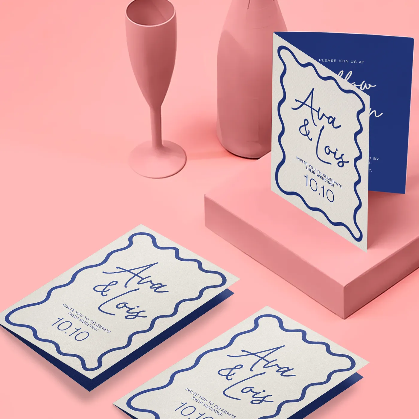Three folded wedding invitations for "Ava & Lois" feature a wavy navy blue border and script font on a light pink surface, with one invitation standing upright to show the navy blue interior against a background of a pink wine glass and bottle.
