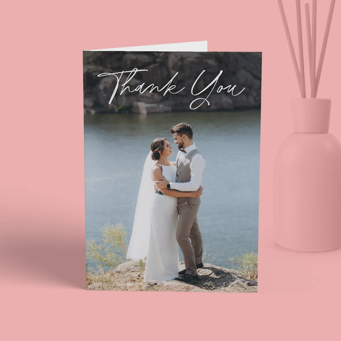 A folded wedding thank-you card stands upright on a light pink background, featuring a full-bleed photo of a bride and groom embracing on a rocky shore by the water. The words "Thank You" are written in a white script font at the top of the photo, and a pink vase with decorative reeds sits to the right of the card.