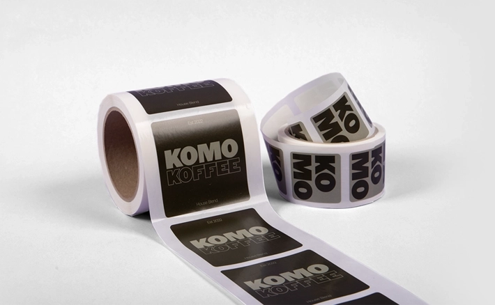 Two rolls of square stickers are shown, featuring a black and grey design for "KOMO KOFFEE" with "House Blend" printed at the bottom.