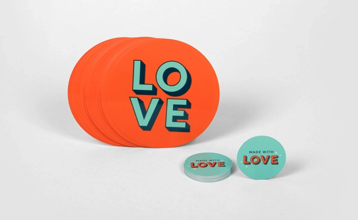 A collection of round individual stickers features a large orange design with "LOVE" in teal block letters and smaller teal stickers that say "MADE WITH LOVE."