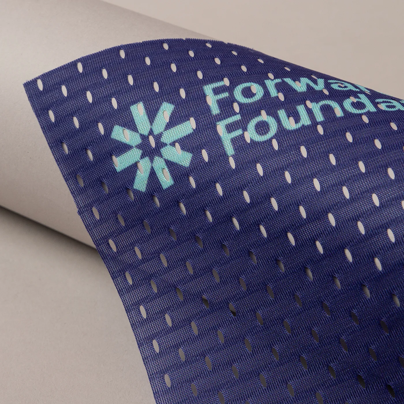 A close-up detail of a dark navy blue mesh banner with a distinct perforated texture. The light teal "Forwards Foundation" logo, featuring a stylized asterisk symbol, is printed clearly on the mesh. The banner is shown partially curved against a smooth, light grey background.