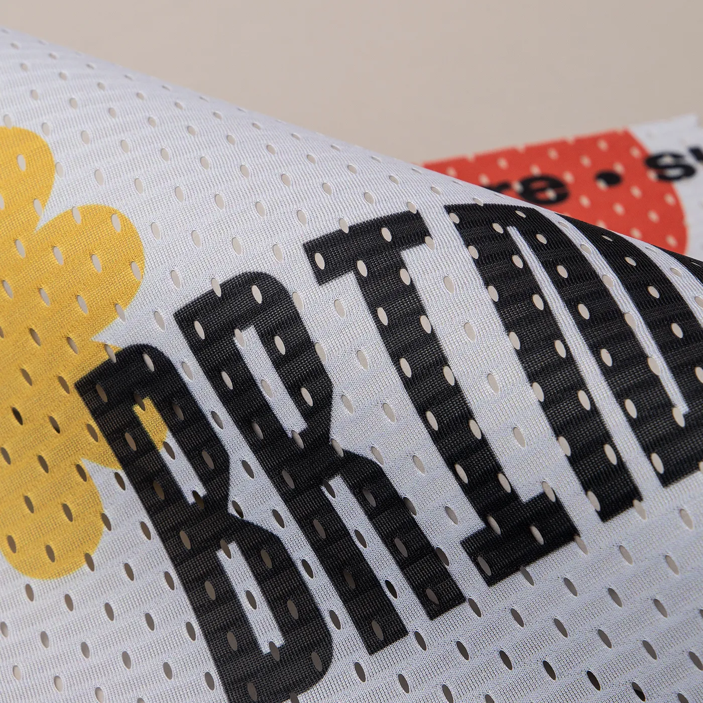 A close-up, angled view showing the texture of a white mesh banner. The material features a pattern of small, oval-shaped perforations. Large, bold black letters and a portion of a yellow graphic are printed across the mesh surface. In the background, a second layer of the banner is visible with a red graphic.