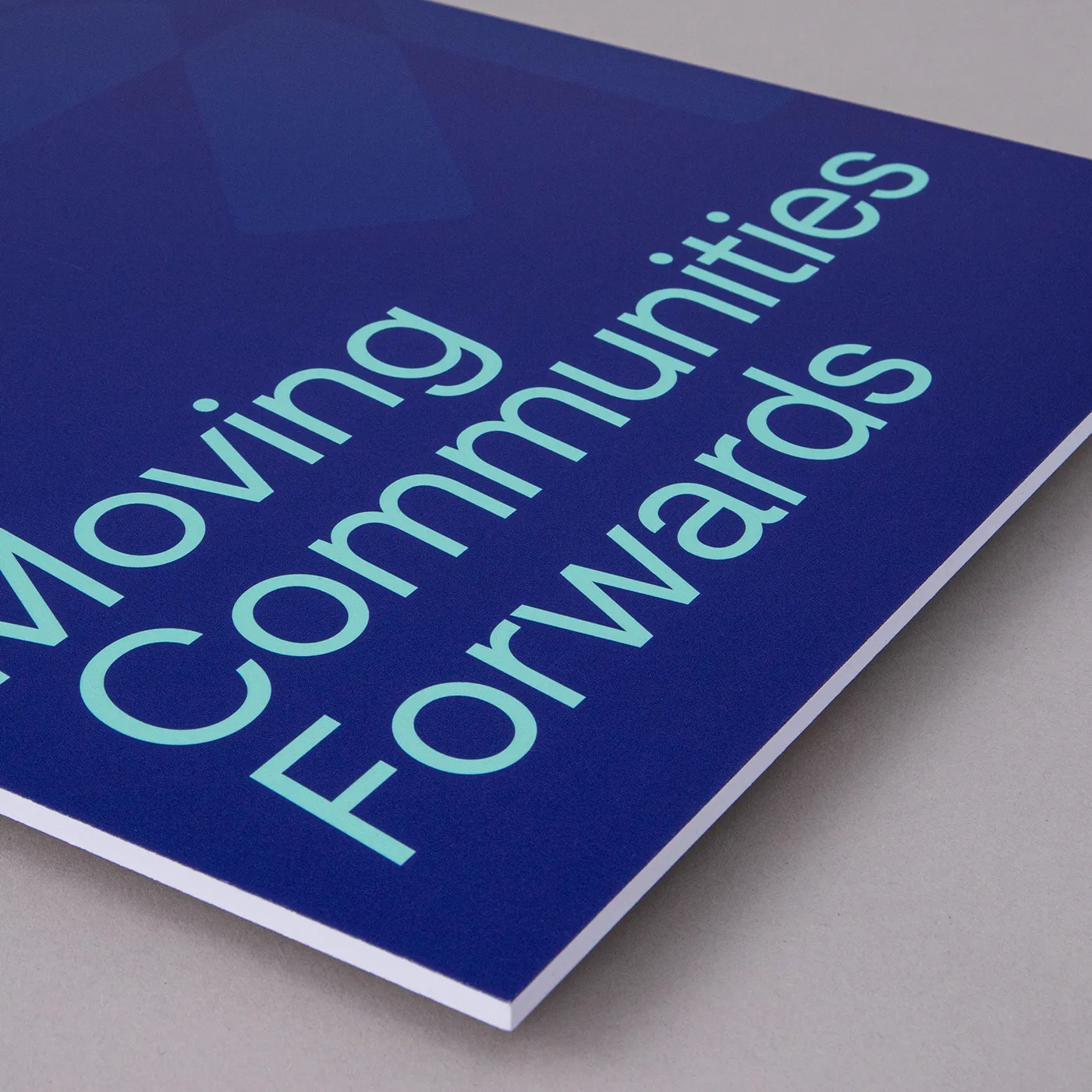 A high-angle close-up of a dark blue sign with light teal text that reads "Moving Communities Forwards." The text is arranged diagonally in a bold, sans-serif font. The sign is made of a thick white material with a blue printed surface and is resting on a neutral grey background.