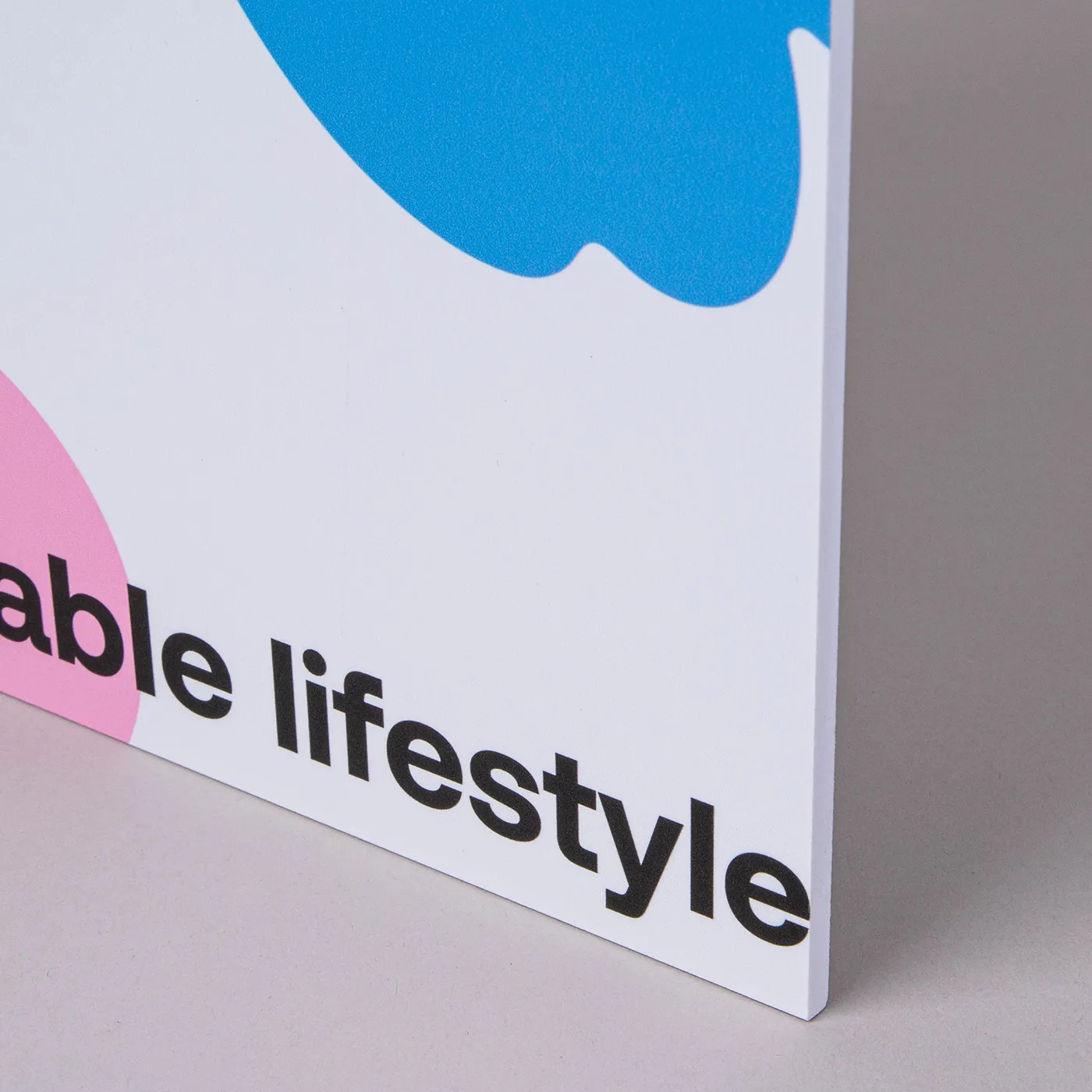 An extreme close-up of the edge of a white rigid sign. The words "able lifestyle" are visible in bold black lowercase text at the bottom. Above the text is a portion of a bright blue abstract shape and a small section of a pink shape on the left edge. The sign is shown from a side angle, highlighting its thickness.
