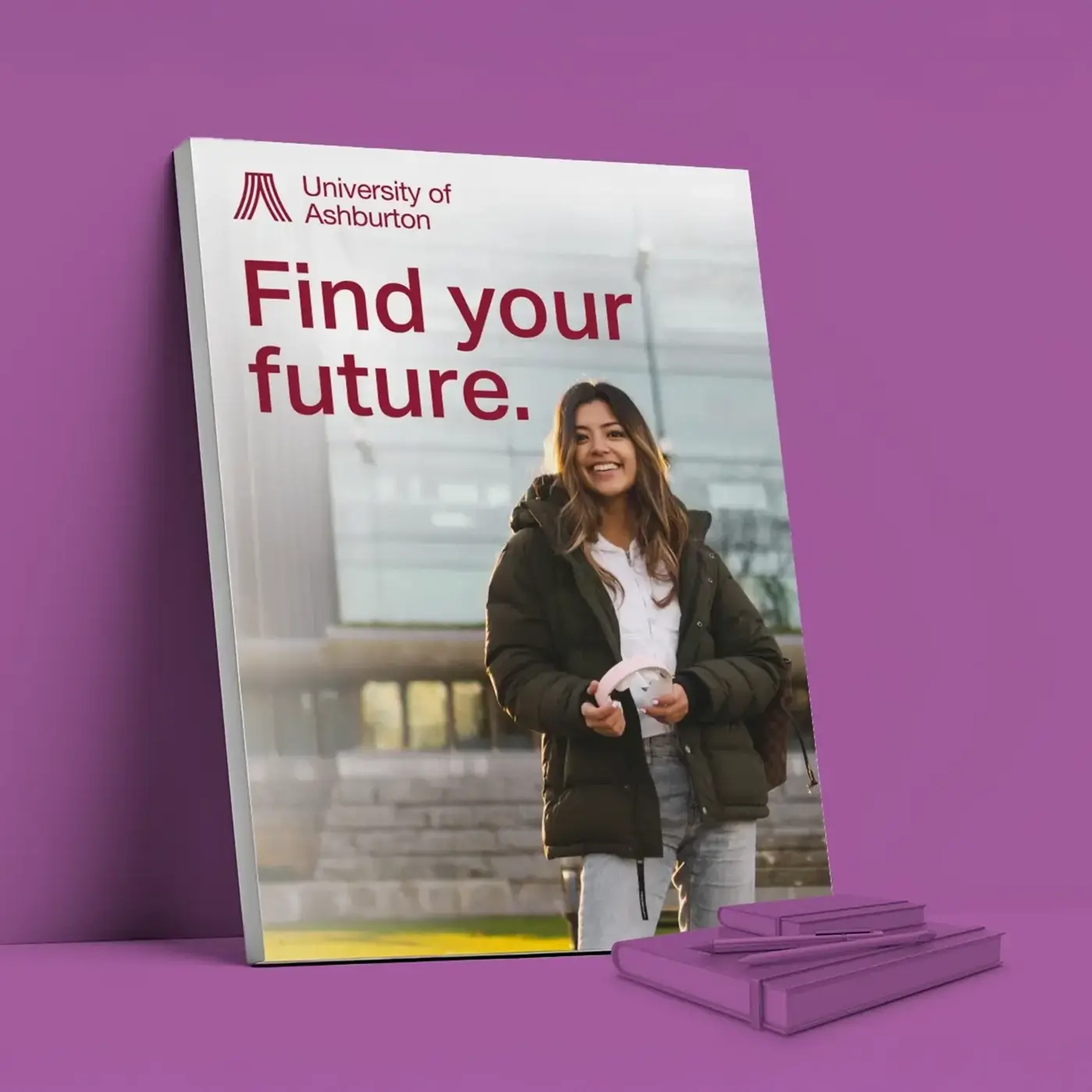 A rectangular display board for the "University of Ashburton" leans against a solid purple wall. The board features a photograph of a smiling student wearing a puffer jacket and holding pink headphones, with a campus building in the background. The text "Find your future." is printed in large, maroon letters at the top. On the purple floor in front of the board, a stack of purple notebooks and a purple pen are arranged.