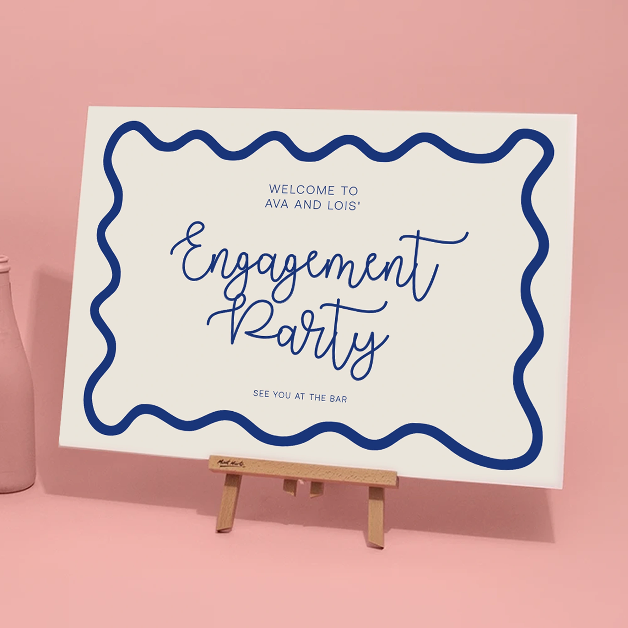 A horizontal engagement party welcome sign for "Ava and Lois" displayed on a small wooden tabletop easel against a solid pink background. The white sign features a thick, wavy navy blue border. The words "Engagement Party" are written in a large, elegant navy blue script, flanked by the phrases "WELCOME TO AVA AND LOIS'" above and "SEE YOU AT THE BAR" below in a simple sans-serif font. A matte pink bottle is partially visible on the left.