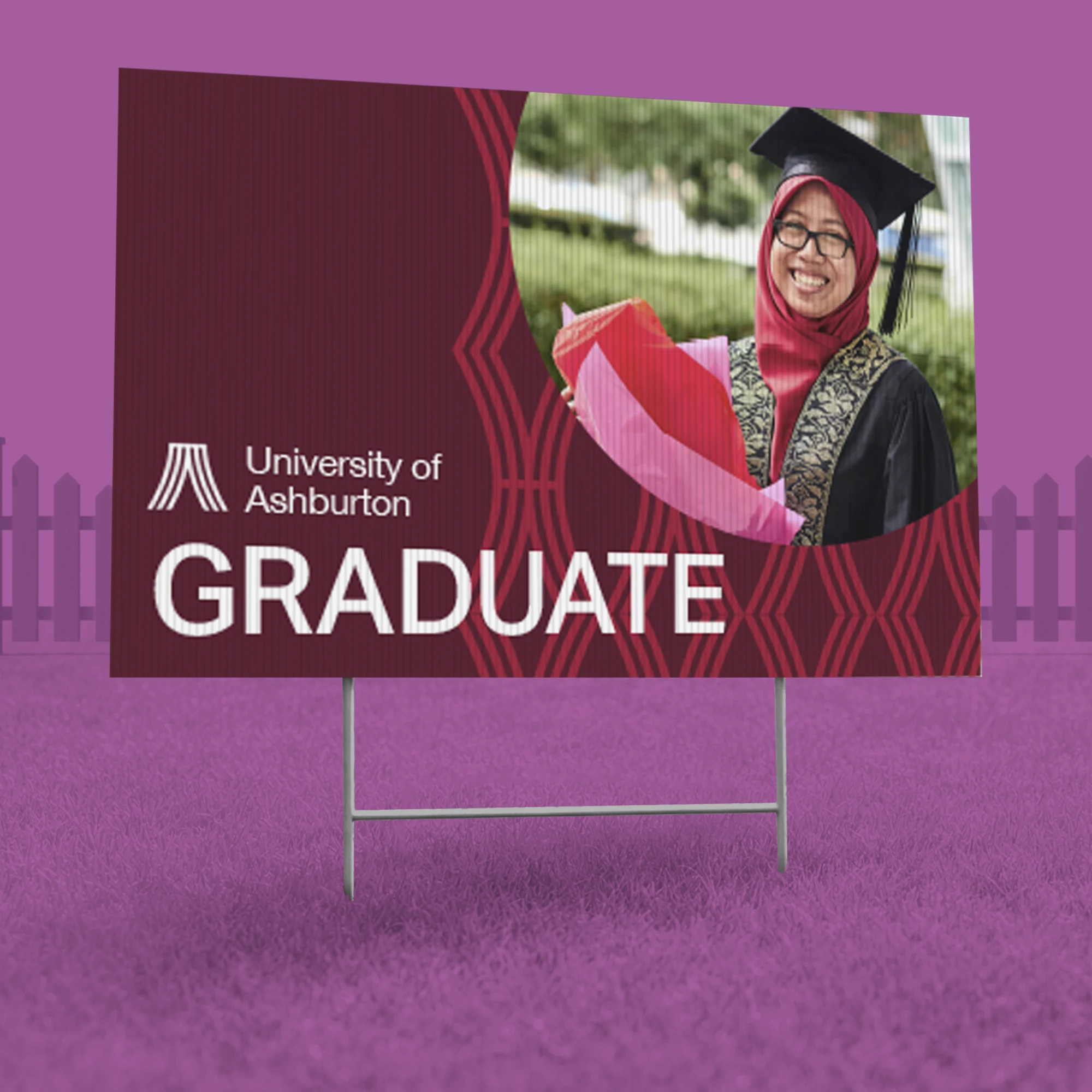 A "University of Ashburton" graduation yard sign displayed on a metal H-stake. The sign has a deep maroon background with geometric patterns and large white text that reads "GRADUATE." It features a photo of a smiling female graduate in a cap and gown holding a bouquet. The entire scene is set against a vibrant purple grass and fence background.