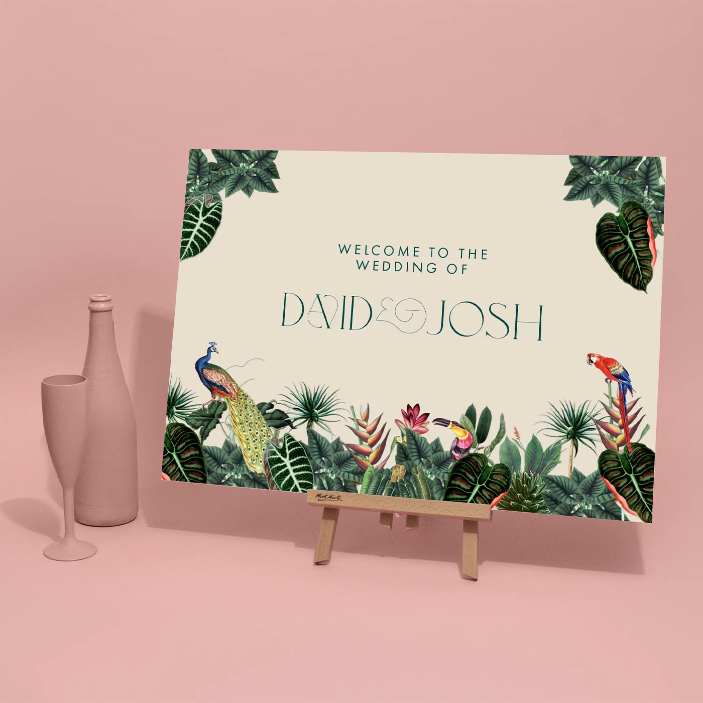 A tropical-themed wedding welcome sign for "David & Josh" displayed on a small wooden tabletop easel against a solid pink background. The sign features lush botanical illustrations, including large green leaves, a peacock, and a parrot along the bottom and corners. To the left of the easel, a matte pink champagne flute and bottle are positioned as decorative props.