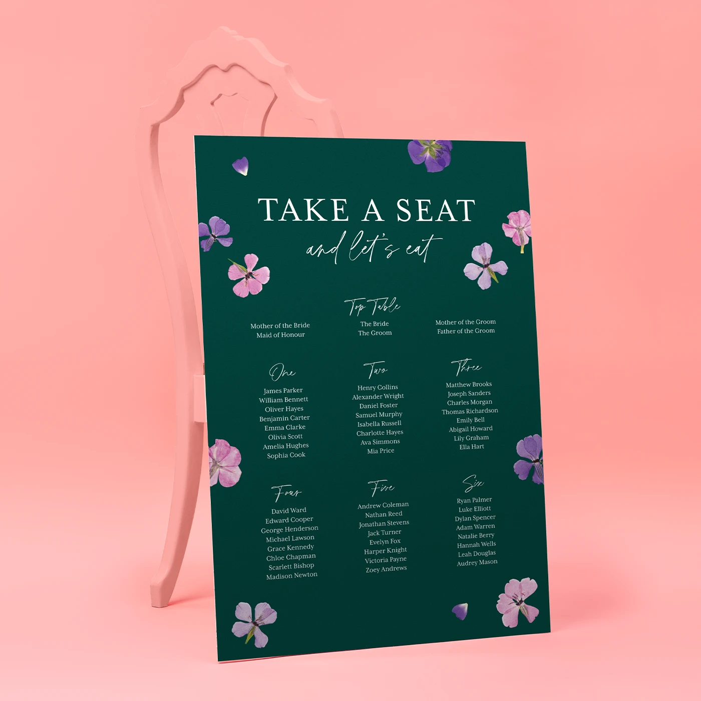 A large, dark forest green wedding seating chart leaning against a light pink ornamental chair. The sign is titled "TAKE A SEAT and let's eat" in white serif and script fonts, decorated with scattered pressed purple and pink flowers. It lists names for a "Top Table" and tables one through six.
