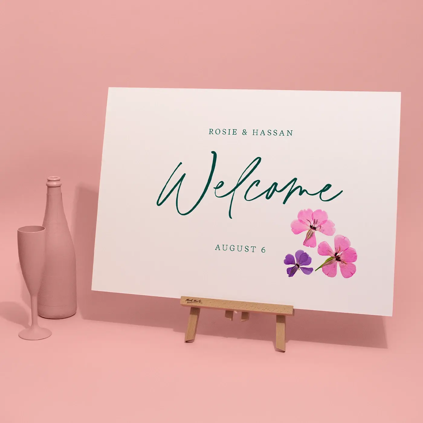 A white wedding welcome sign featuring the names "Rosie & Hassan" and the date "August 6" is displayed on a small wooden easel against a pink background, decorated with green script and pressed pink and purple flowers.