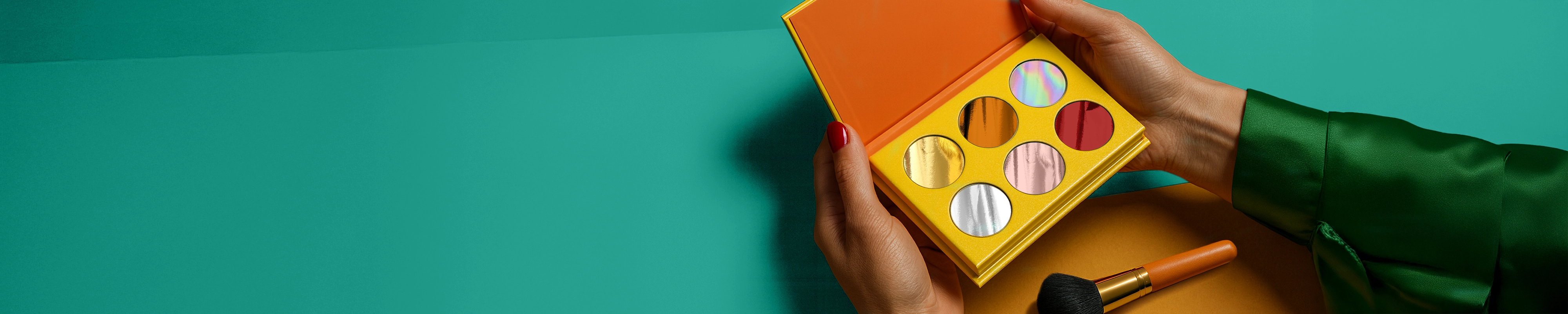 A wide banner showing hands holding an open yellow palette with six circular metallic and holographic foil samples inside. A makeup brush rests below on a dual-toned teal and gold background, with ample copy space on the left.