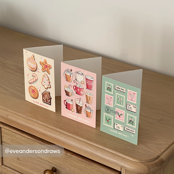 Three personalized Christmas Cards displaying illustrations of holiday cookies, cozy warm drinks, and postage stamps displayed on a wooden surface.